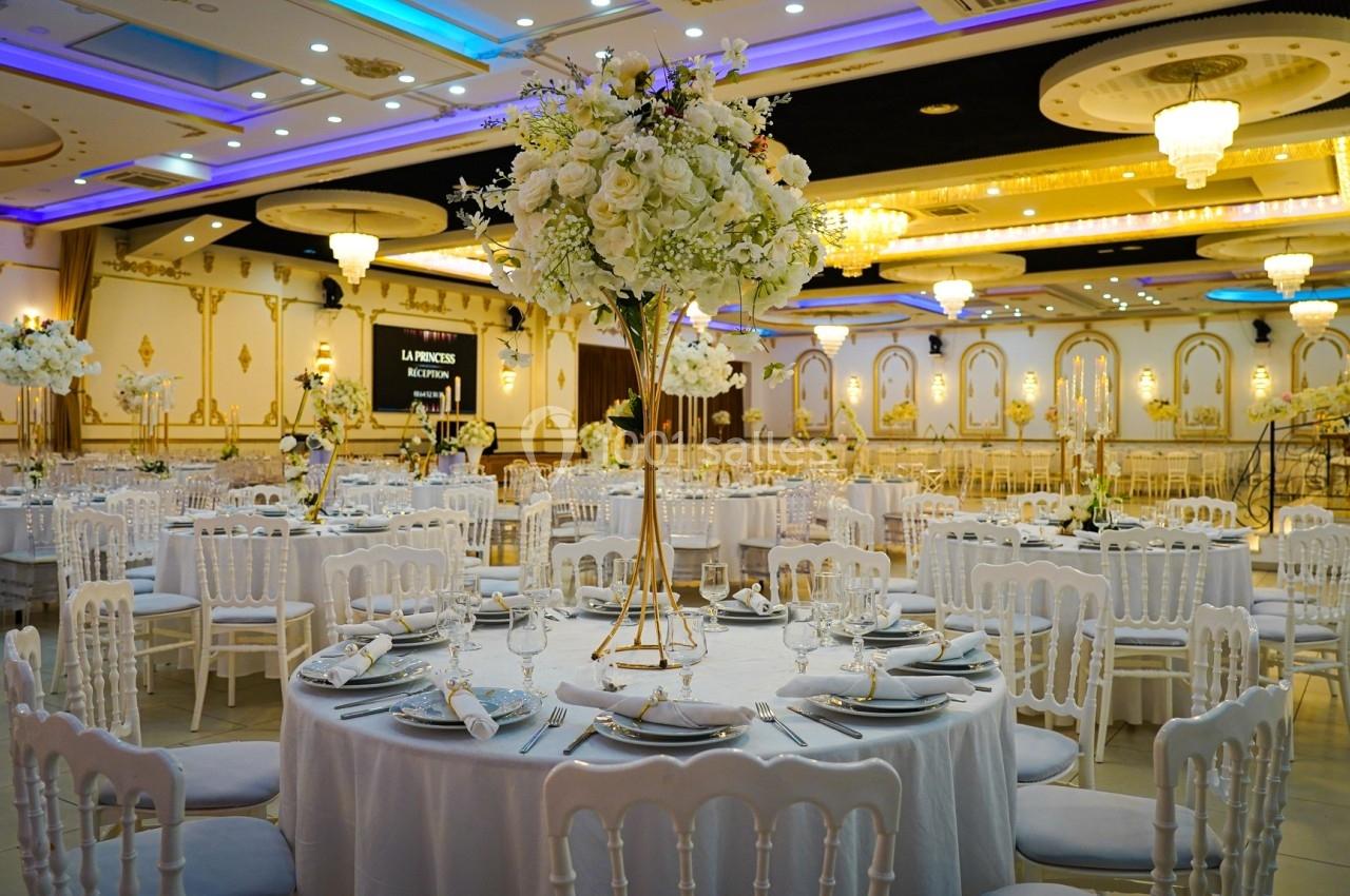 Salle de réception élégante avec tables rondes dressées, décorations florales et lustres suspendus.