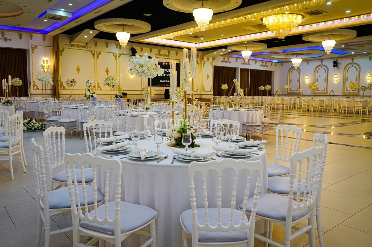 Salle de réception élégante avec tables rondes dressées, chaises blanches et décorations lumineuses.