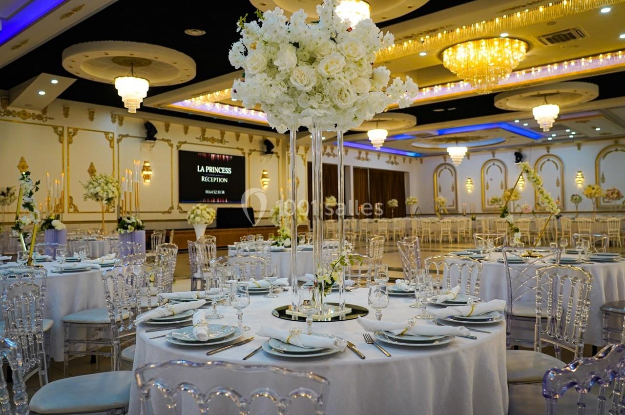 Salle de réception élégante avec tables rondes dressées, chaises transparentes et décorations florales blanches.
