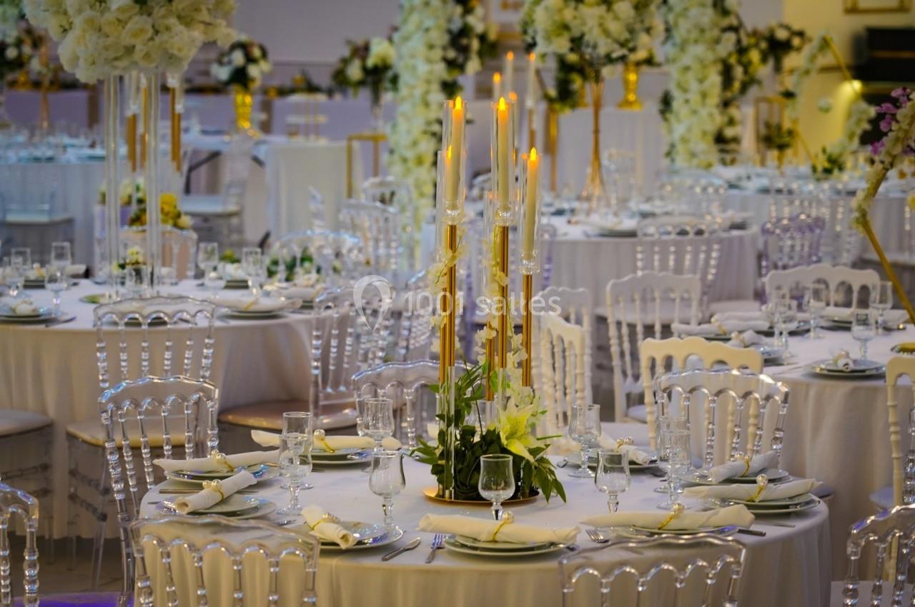 Salle de réception décorée avec des tables rondes, nappes blanches, chandeliers dorés et arrangements floraux.