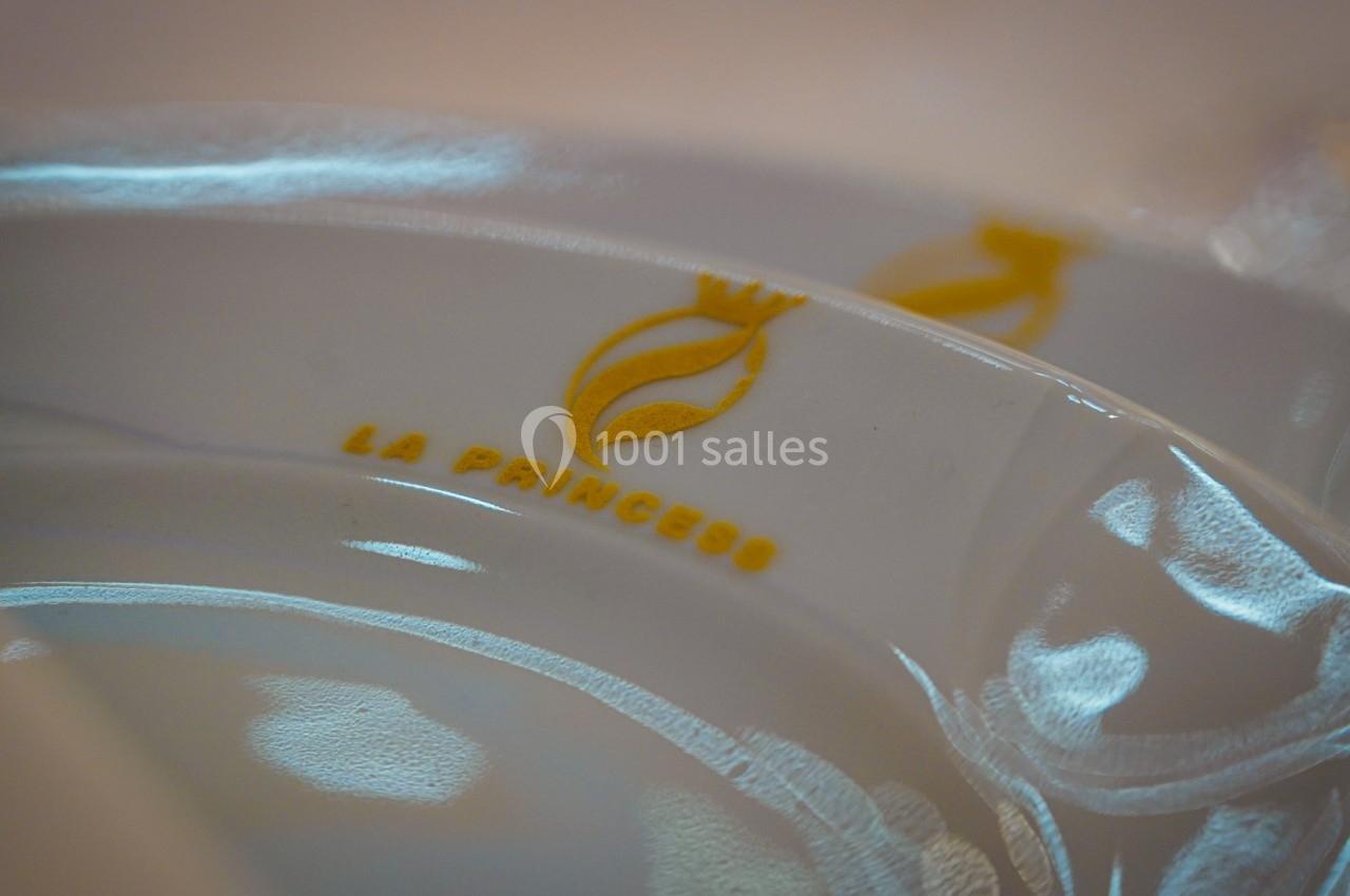 Assiette blanche avec un logo doré représentant une couronne et une flamme, accompagnée du texte ’La Princess’.