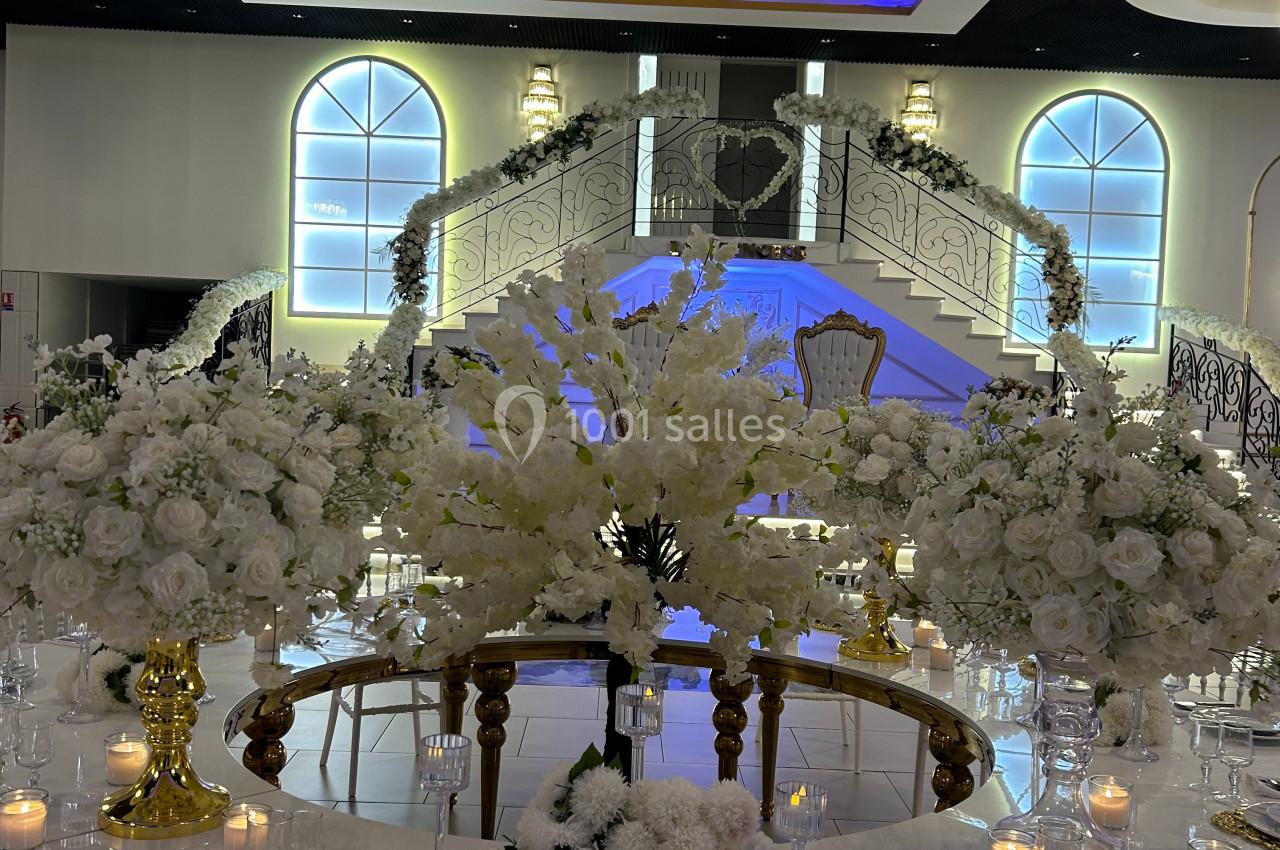 Salle de réception élégante avec table décorée de fleurs blanches, chandelles et escalier illuminé en arrière-plan.