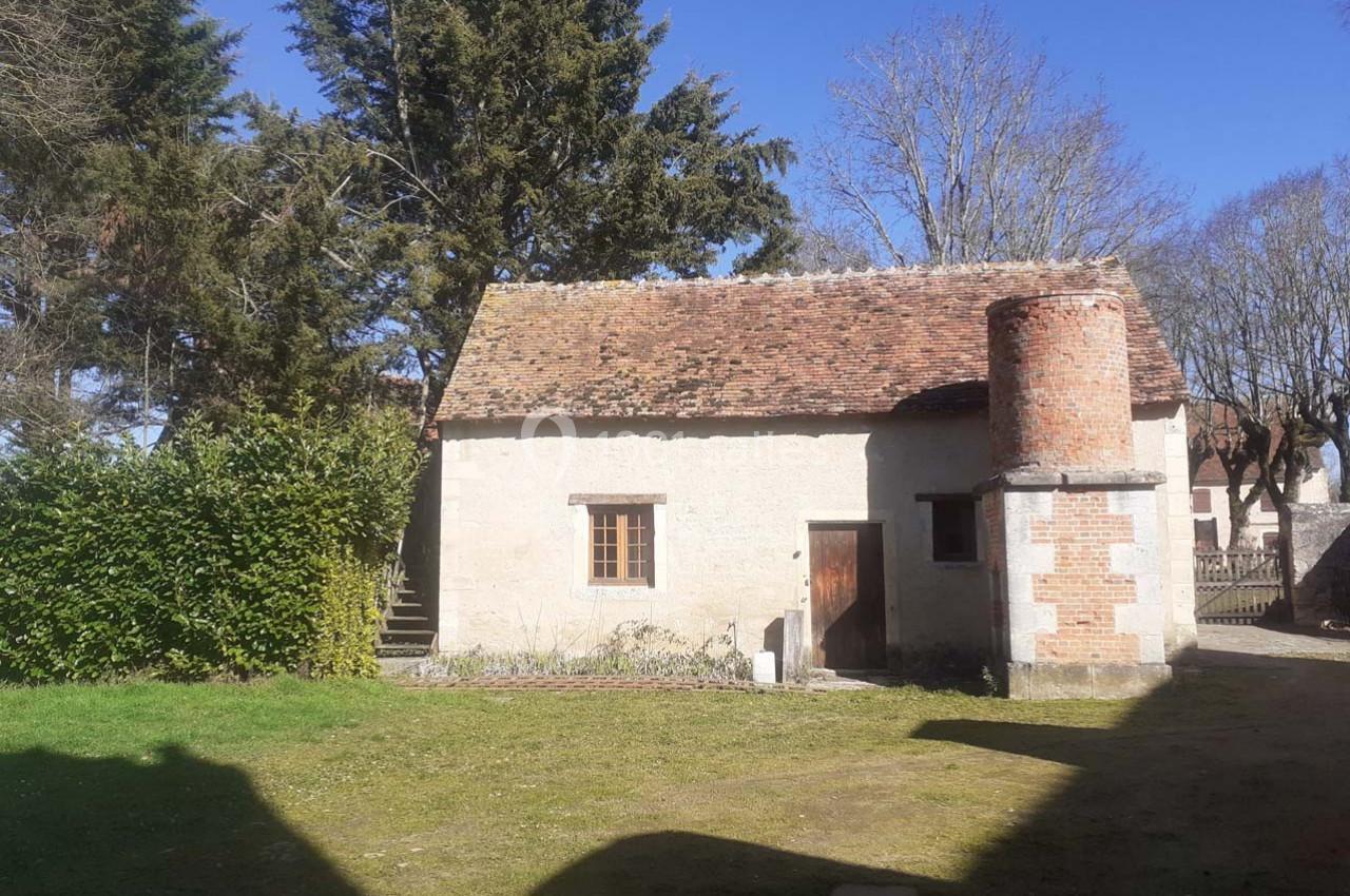 Petite maison en pierre avec un toit en tuiles, une cheminée en briques et entourée d'arbres et de pelouse.