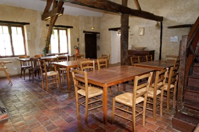 Salle rustique avec tables et chaises en bois, sol en carrelage ancien, poutres apparentes et lumière naturelle.