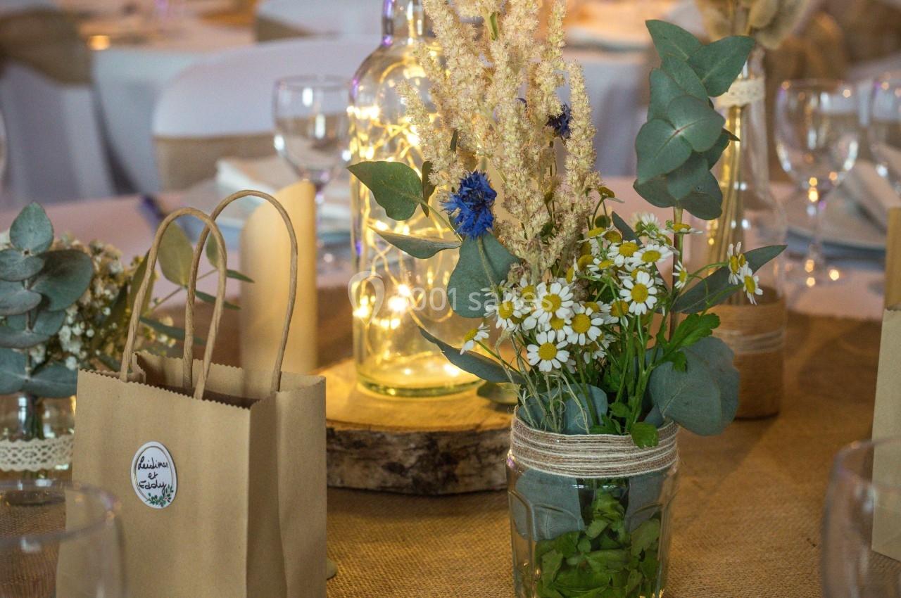 Centre de table avec fleurs séchées, marguerites et feuillage dans des bocaux en verre, décoré de guirlandes lumineuses.