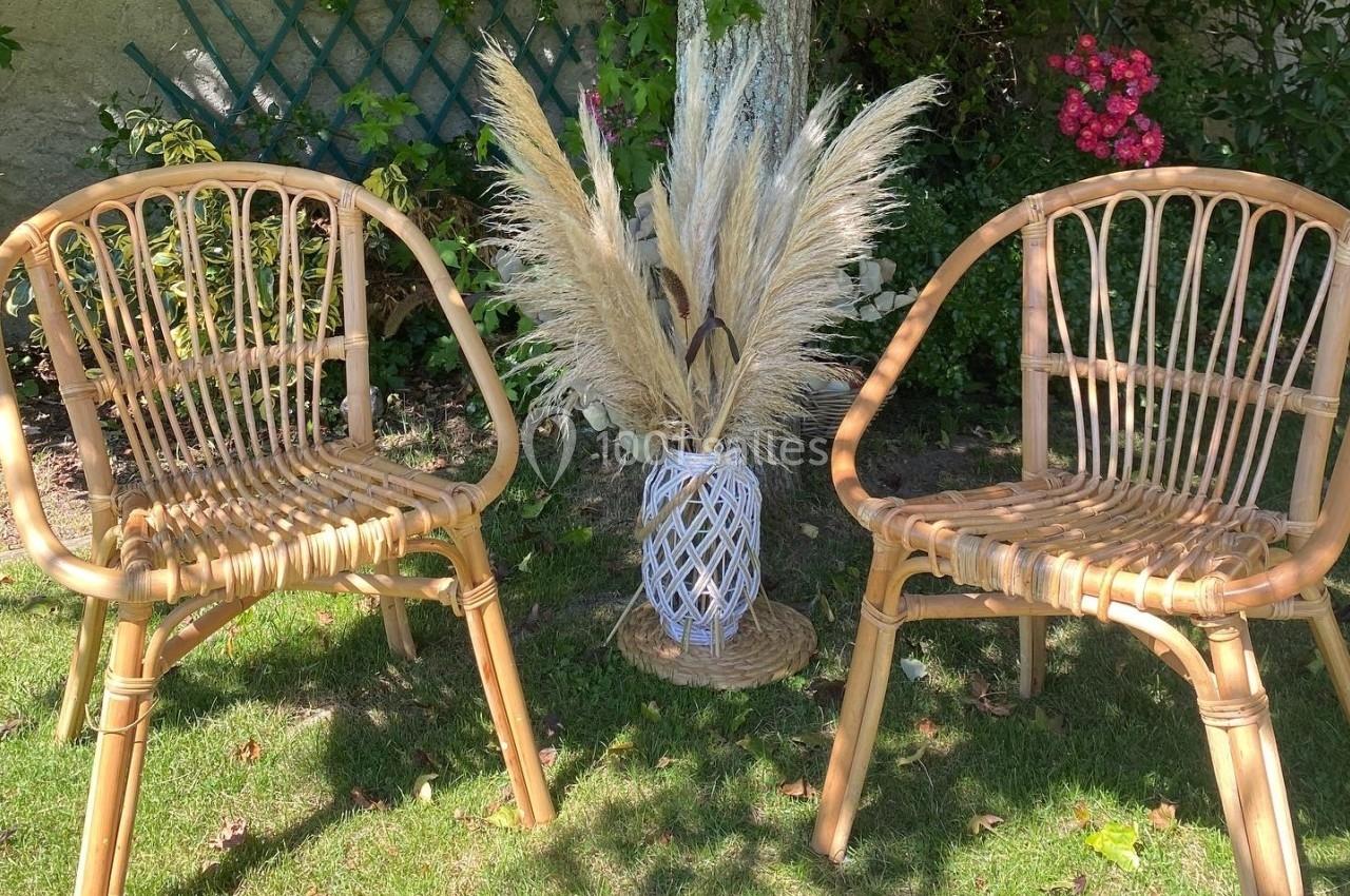 Deux fauteuils en rotin disposés sur une pelouse, encadrant un vase en osier rempli de pampas, avec un fond de jardin fleuri.