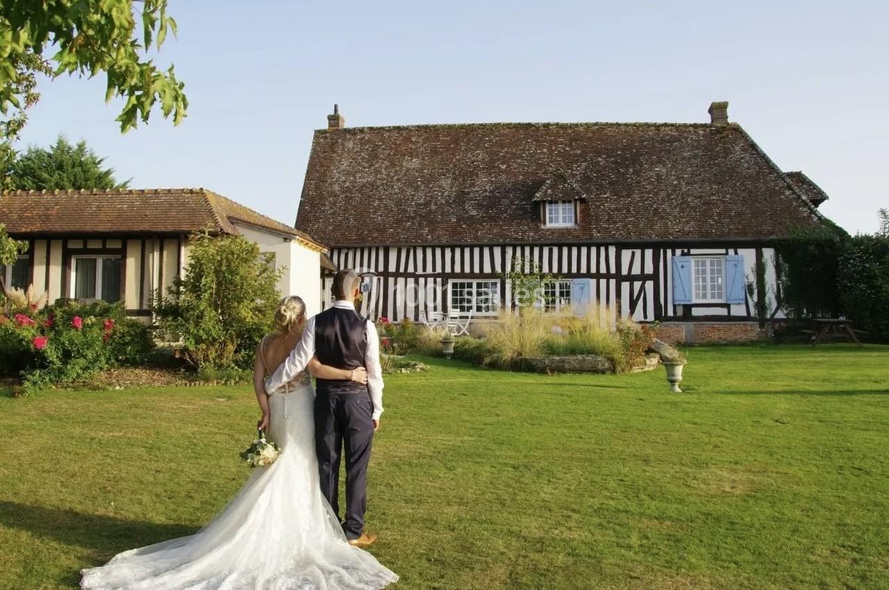 Un couple en tenue de mariage regarde une maison à colombages dans un jardin verdoyant par une journée ensoleillée.
