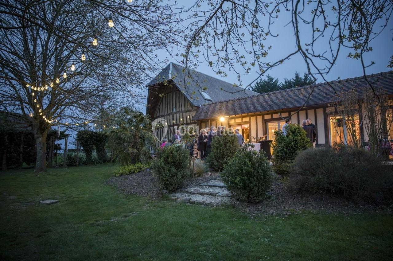 Maison à colombages éclairée en soirée, entourée d'arbres et de pelouse, avec des invités rassemblés à l'extérieur.