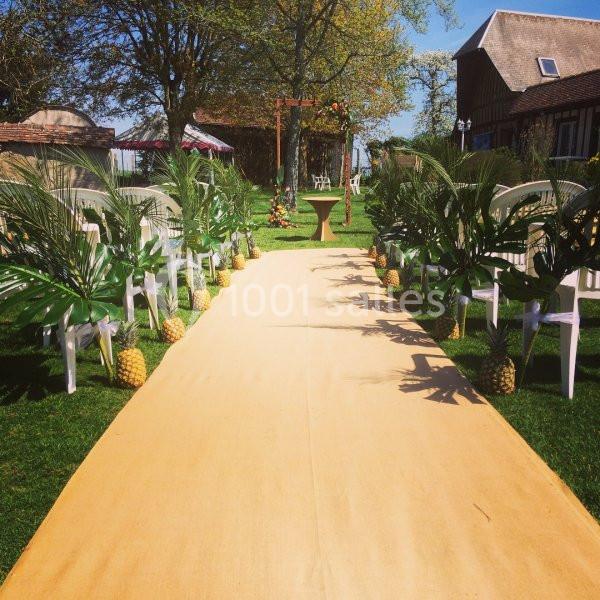 Allée de cérémonie extérieure bordée de feuilles tropicales et d'ananas, avec des chaises blanches disposées de chaque côté.