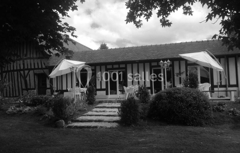 Façade d'une maison à colombages avec terrasse, parasols blancs, mobilier de jardin et végétation environnante.