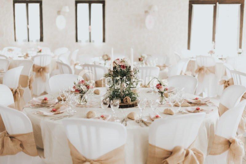 Salle de réception décorée avec des tables rondes, nappes blanches, centres de table fleuris et chaises ornées de rubans.