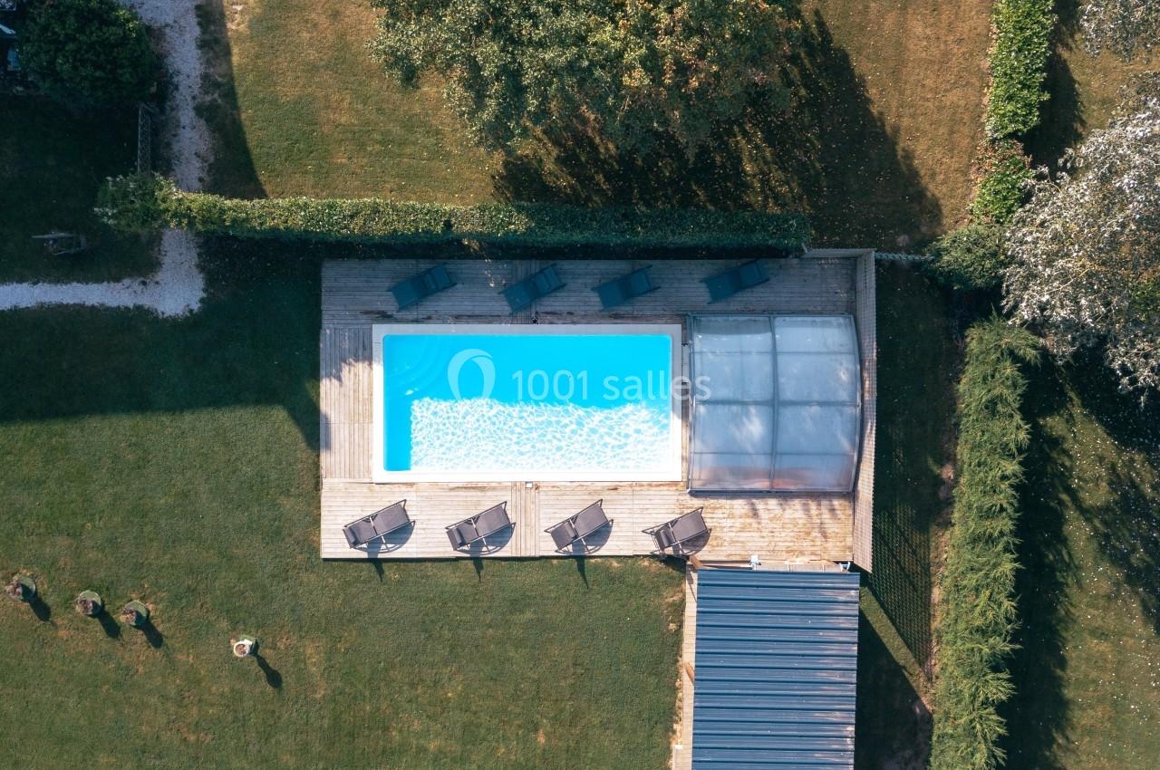 Vue aérienne d'une piscine rectangulaire entourée de transats sur une terrasse en bois, dans un jardin verdoyant.