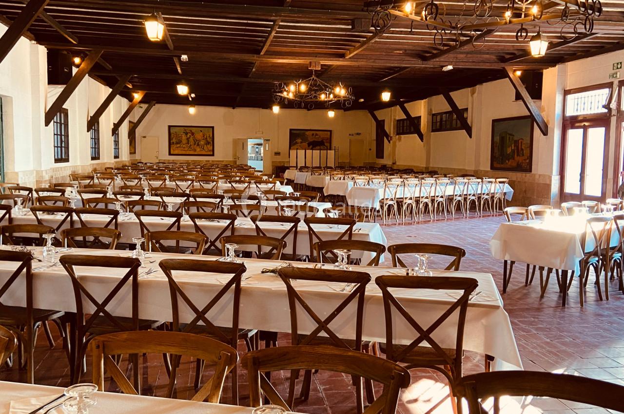 Salle de réception spacieuse avec de longues tables dressées, chaises en bois et éclairage suspendu chaleureux.