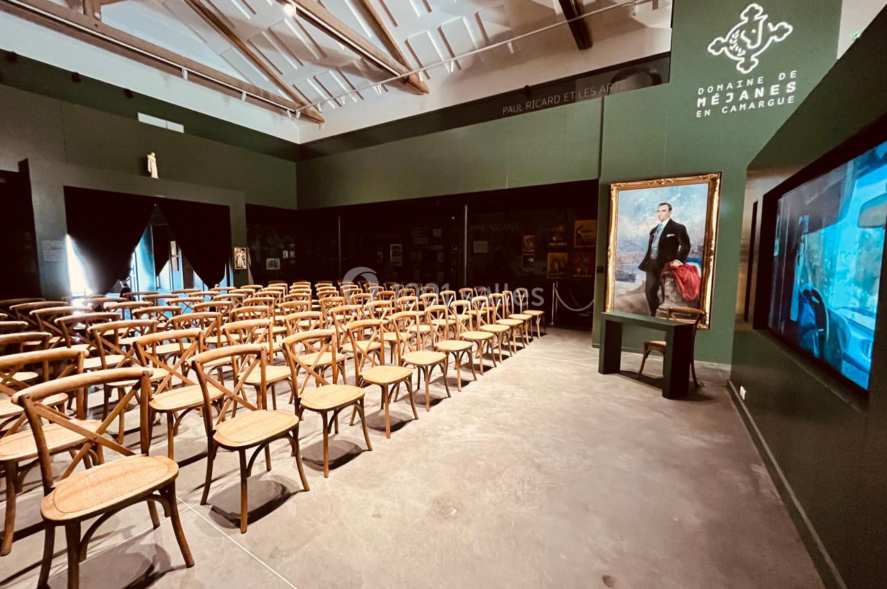 Salle d'exposition avec des rangées de chaises en bois, un tableau portrait et un écran interactif sur la droite.