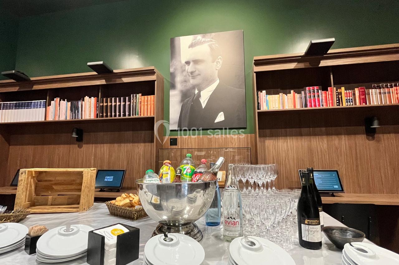 Table dressée avec des assiettes, verres, boissons et viennoiseries devant une bibliothèque et un portrait en noir et blanc.