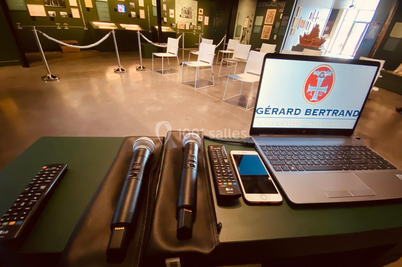 Deux microphones, un ordinateur portable et des télécommandes sur une table, avec une salle d'exposition en arrière-plan.