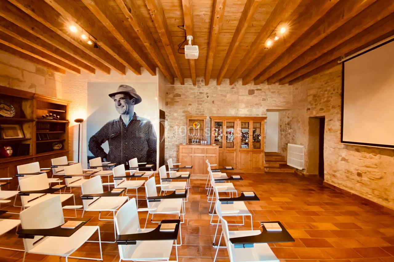 Salle de conférence avec chaises blanches, écran de projection, murs en pierre et photo murale d'une personne souriante.