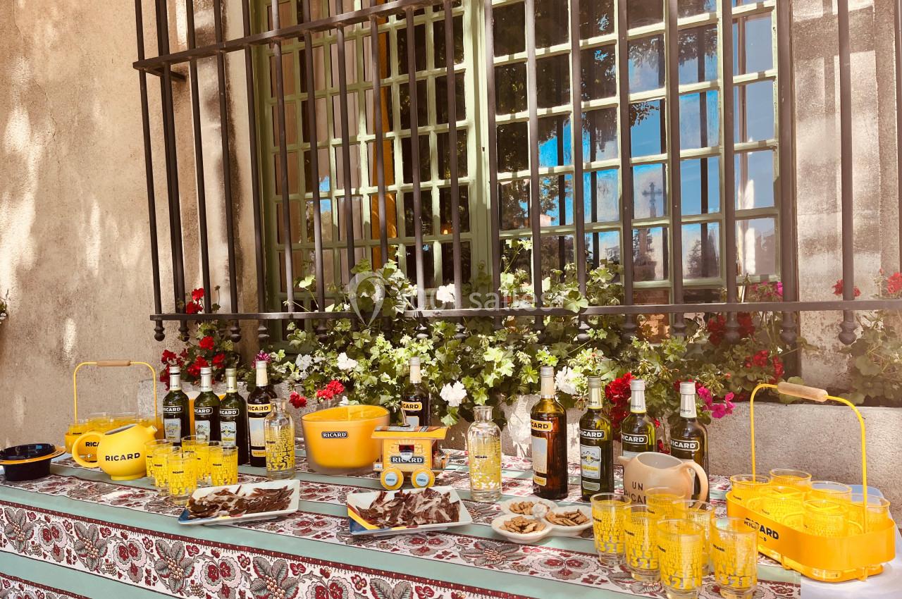 Table dressée en extérieur avec bouteilles, verres, apéritifs et décorations florales devant une fenêtre grillagée.