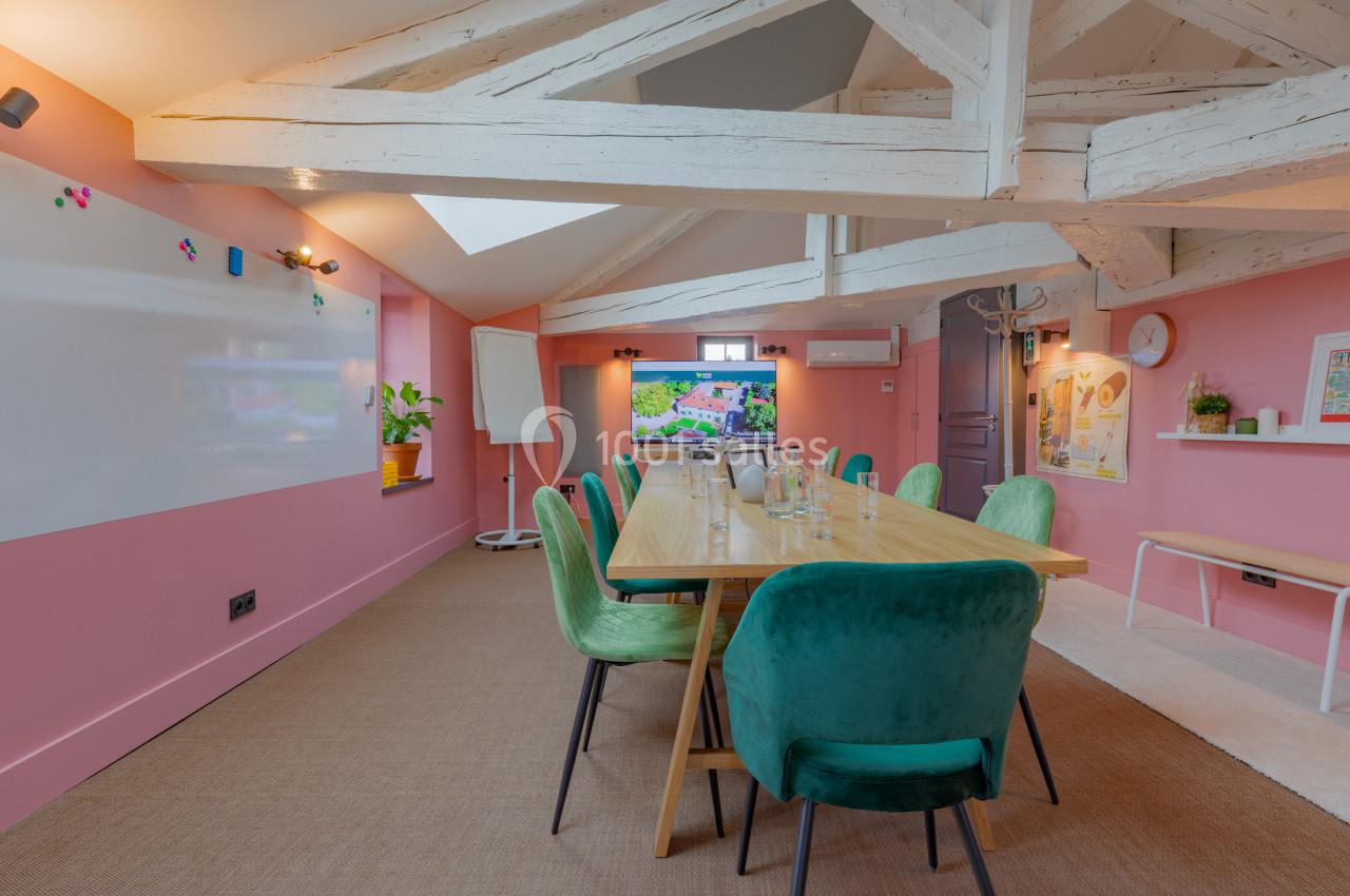 Salle de réunion lumineuse avec murs roses, table en bois entourée de chaises vertes et tableau blanc.