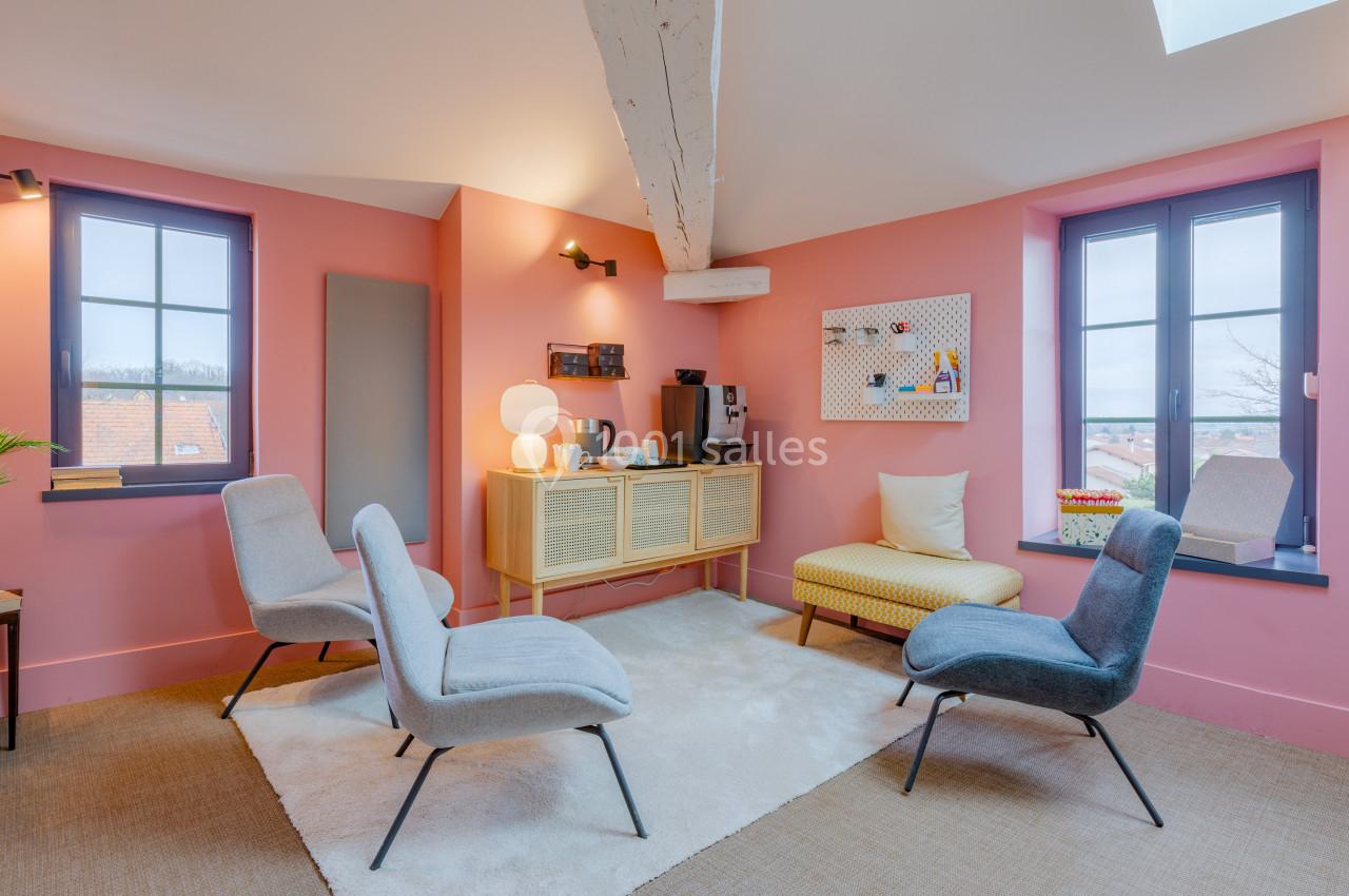 Salle lumineuse avec murs roses, mobilier moderne, fauteuils colorés, tapis clair et vue sur l'extérieur par deux fenêtres.