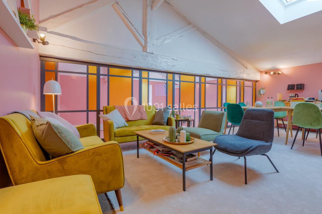 Salon lumineux avec murs roses, mobilier coloré, poutres apparentes et grande verrière laissant passer la lumière naturelle.