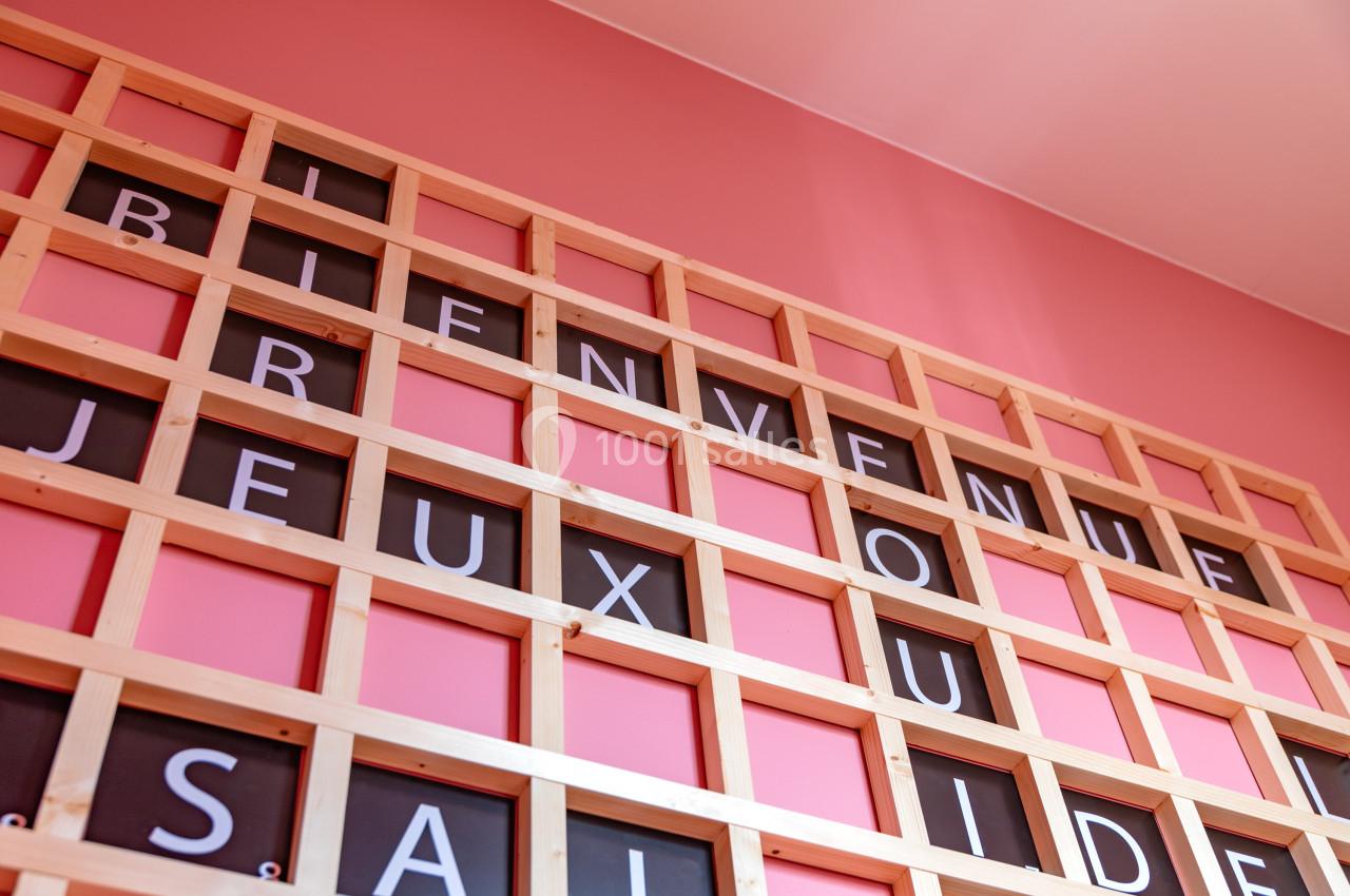 Mur rose avec une grille en bois affichant des lettres formant des mots comme ’Bienvenue’ et ’Jeux’.