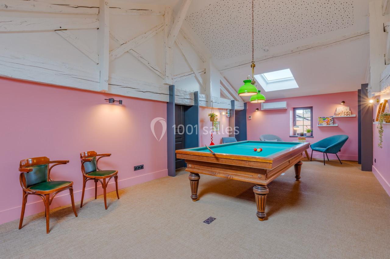 Salle avec un billard en bois, murs roses, chaises vintage, luminaires suspendus et plafond avec poutres apparentes.