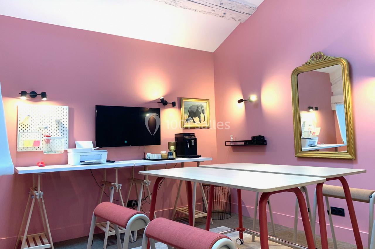 Salle de réunion lumineuse avec murs roses, tables modernes, miroir doré et écran mural.