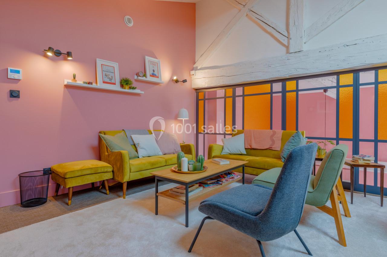 Salon lumineux avec murs roses, mobilier coloré, poutres apparentes et grandes cloisons vitrées aux teintes orangées.