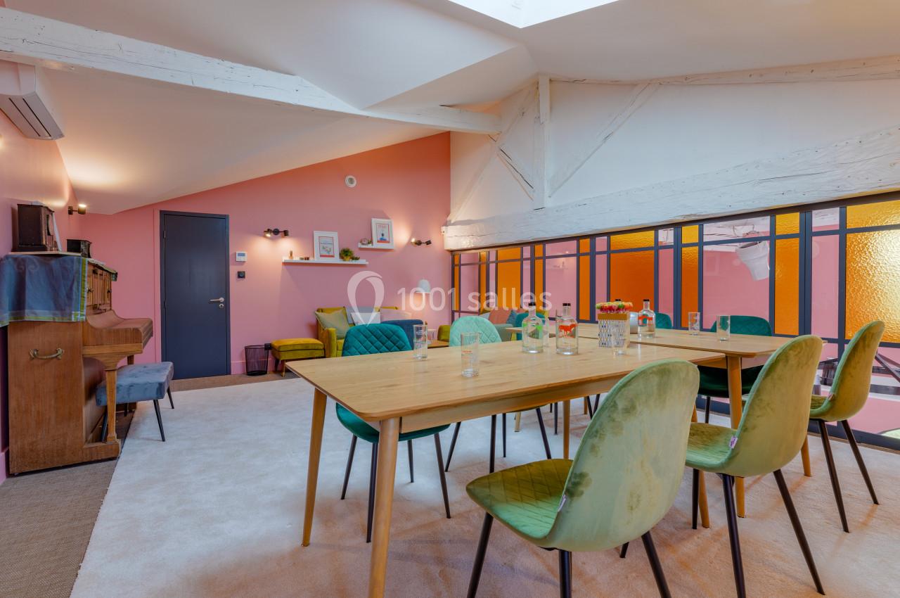Salle lumineuse avec grande table en bois, chaises colorées, murs roses et verrière donnant sur un espace orange.