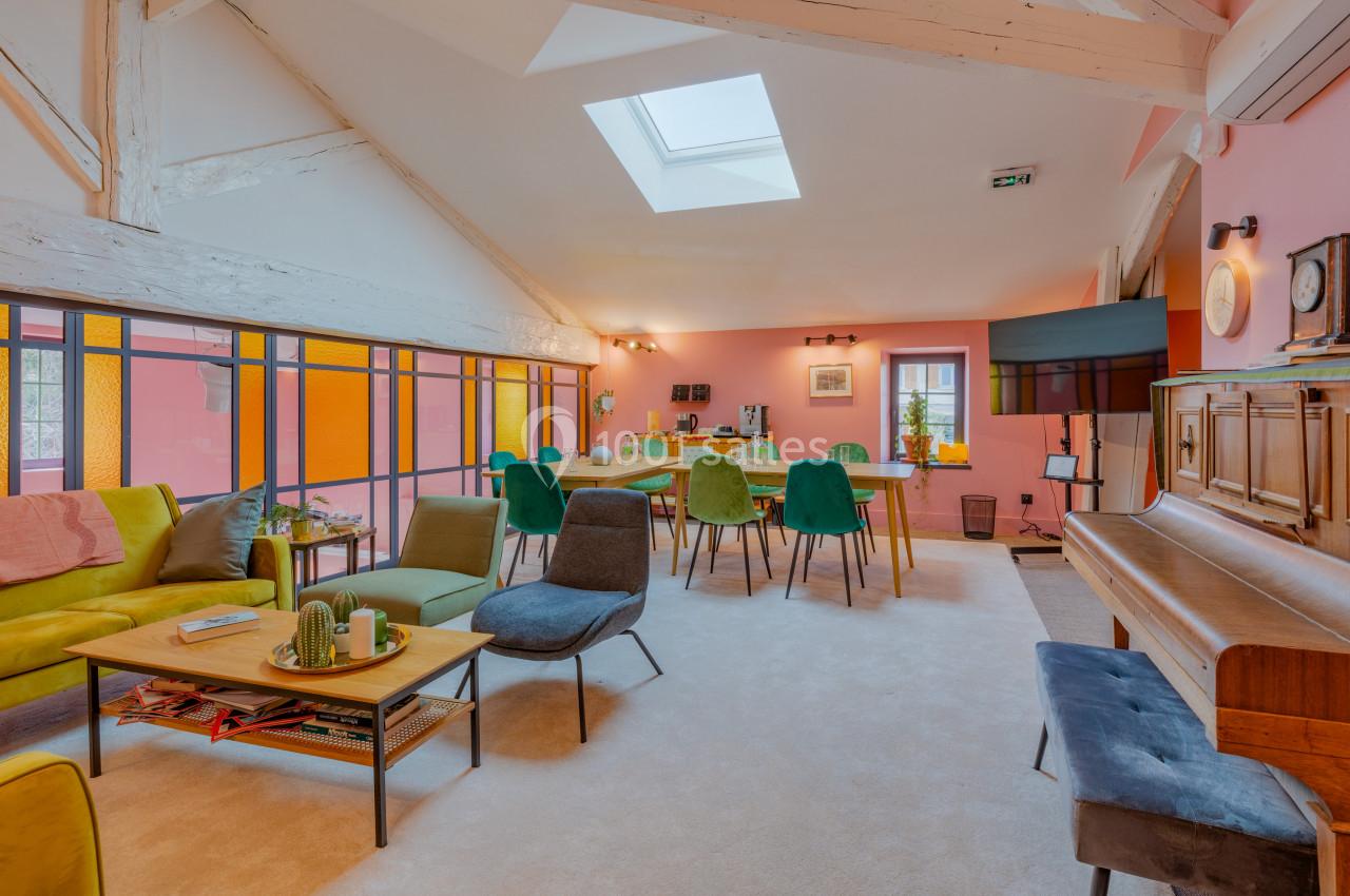 Salle de séjour lumineuse avec murs roses, mobilier coloré, piano, table à manger et grande fenêtre.
