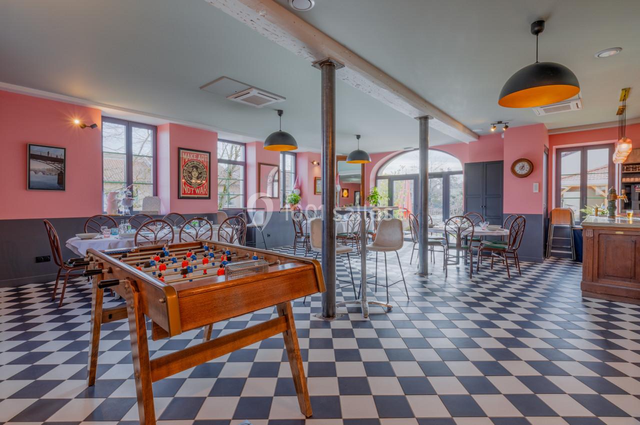 Salle lumineuse avec baby-foot, tables et chaises, sol en damier noir et blanc, murs roses et grandes fenêtres.