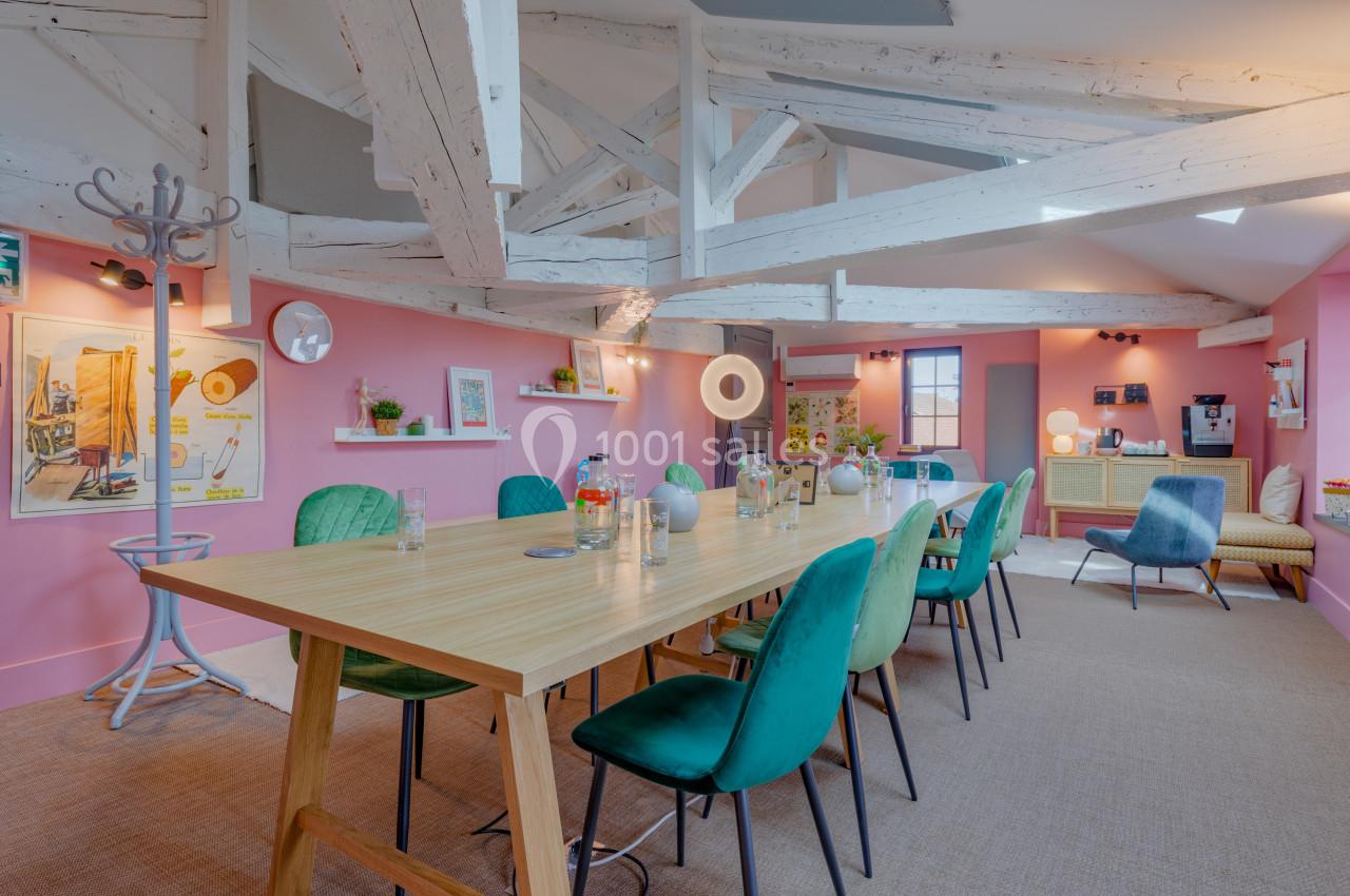 Salle de réunion lumineuse avec murs roses, table en bois, chaises vertes et poutres apparentes au plafond.