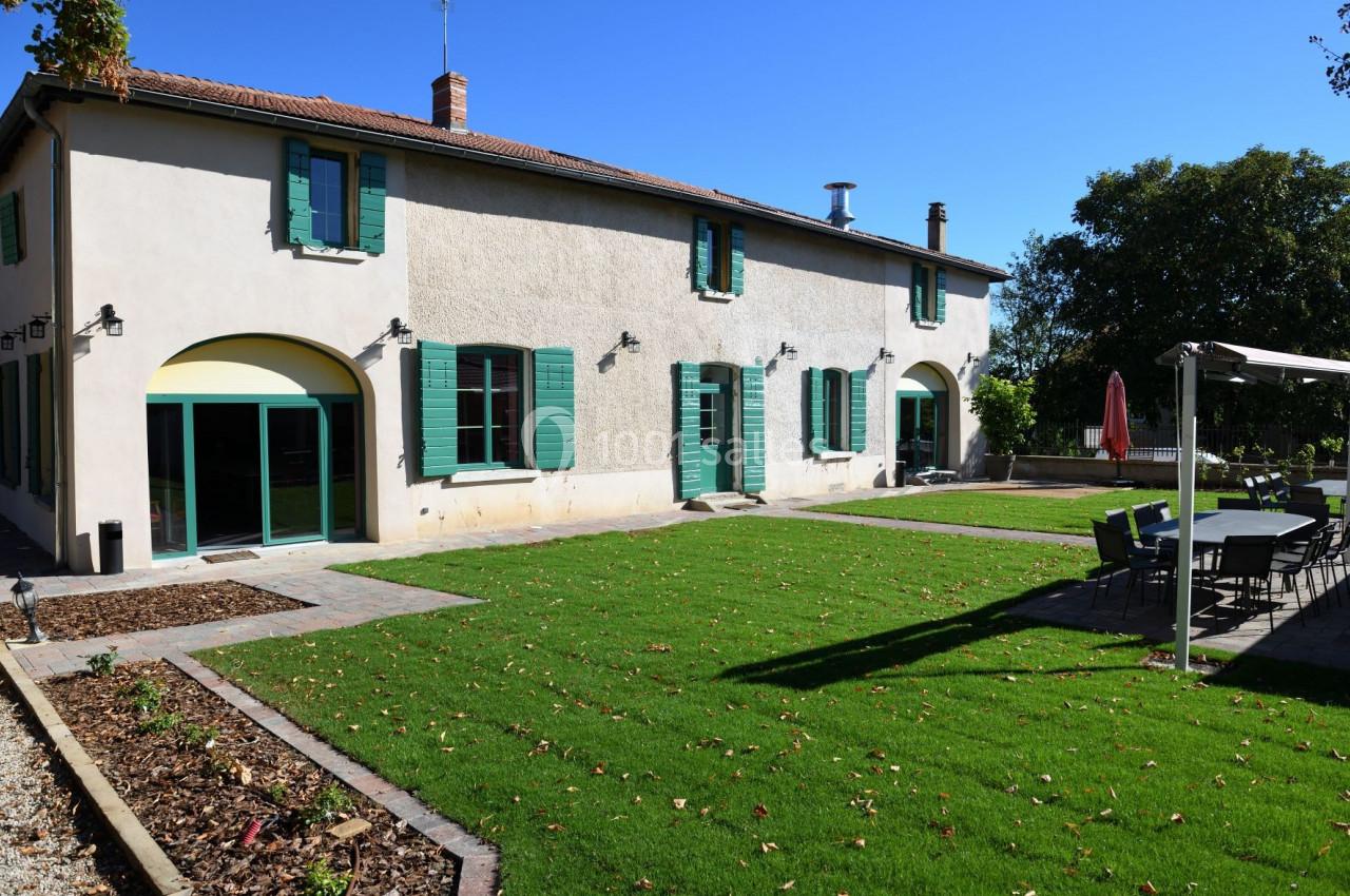 Maison avec façade claire, volets verts, jardin aménagé avec pelouse, terrasse et table extérieure sous un parasol.