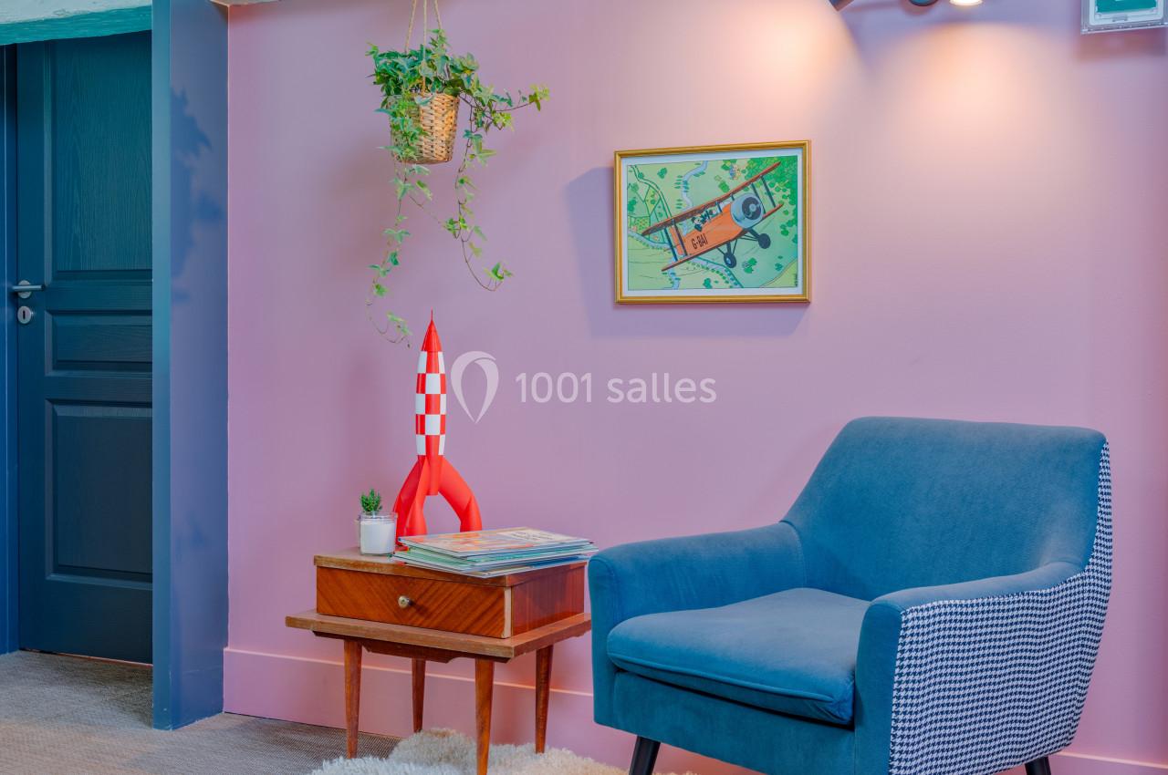 Fauteuil bleu, table en bois avec une fusée miniature, plante suspendue et mur rose décoré d'un tableau.