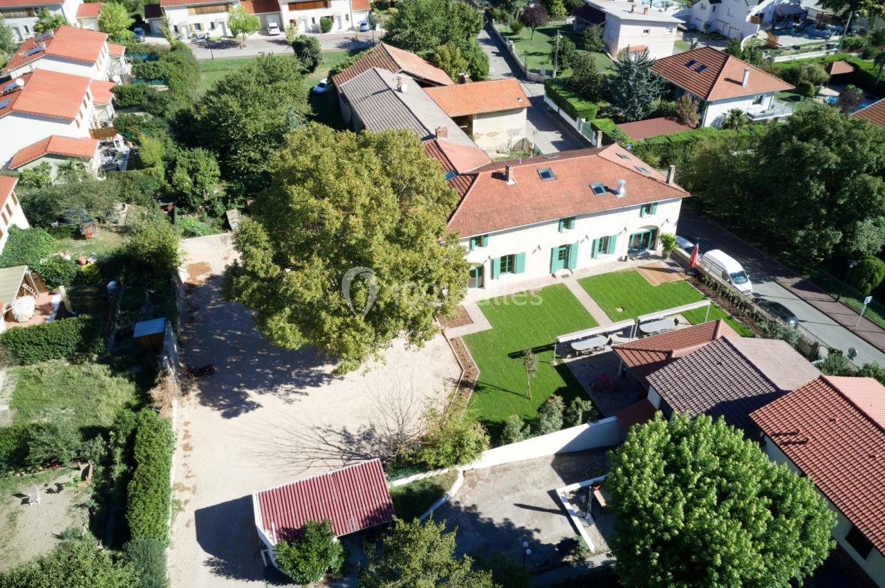 Vue aérienne d'une maison avec jardin, allée gravillonnée, arbres et bâtiments voisins dans un quartier résidentiel.