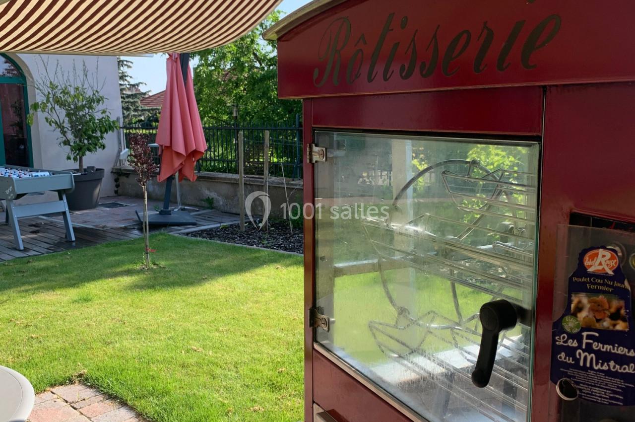 Rôtissoire rouge sous un auvent rayé, installée dans un jardin avec pelouse, terrasse et mobilier extérieur.