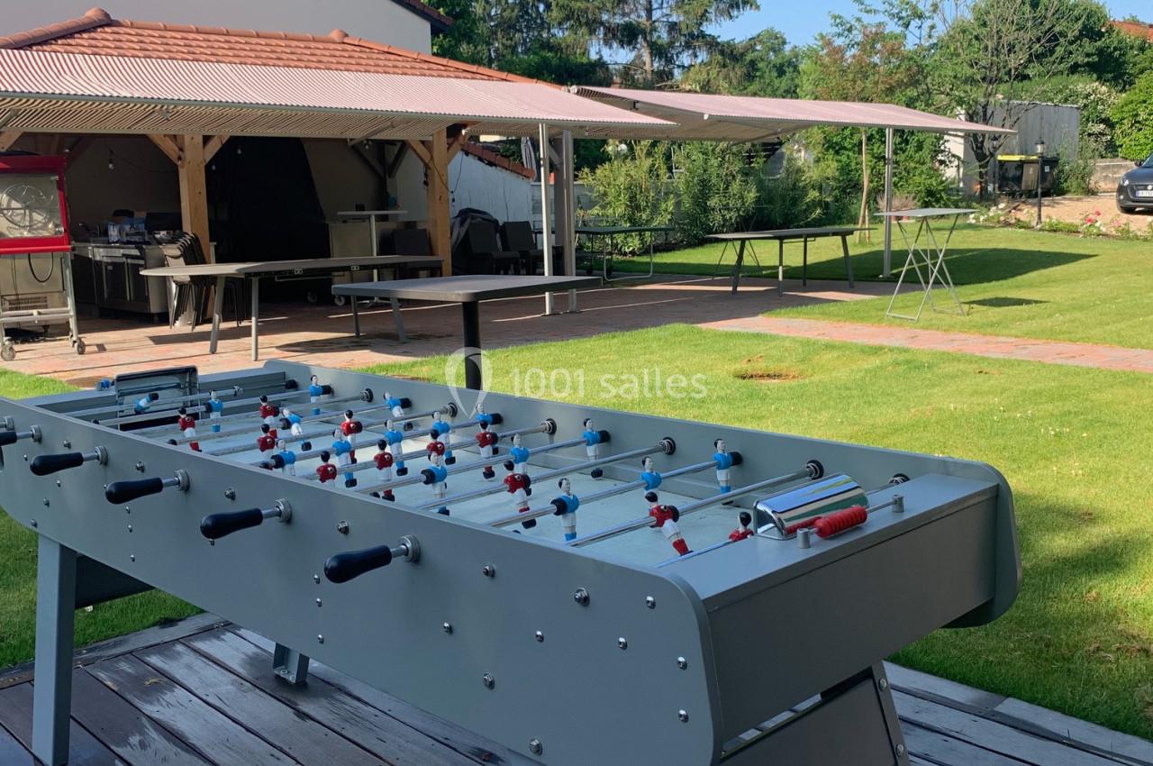 Table de baby-foot en extérieur sur une terrasse en bois, avec un jardin et une pergola en arrière-plan.