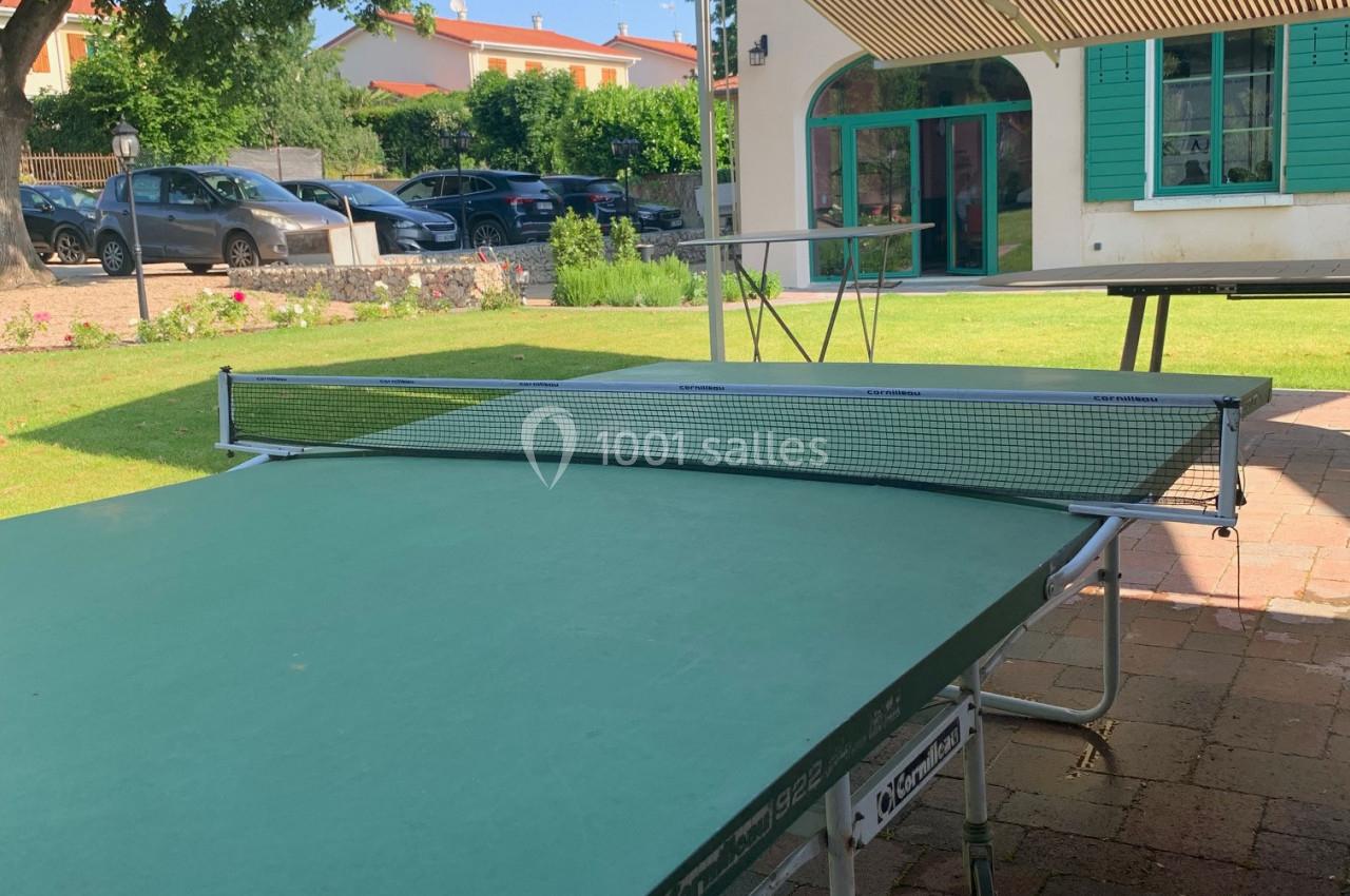 Table de ping-pong verte installée en extérieur sur une terrasse pavée, avec une maison et un jardin en arrière-plan.