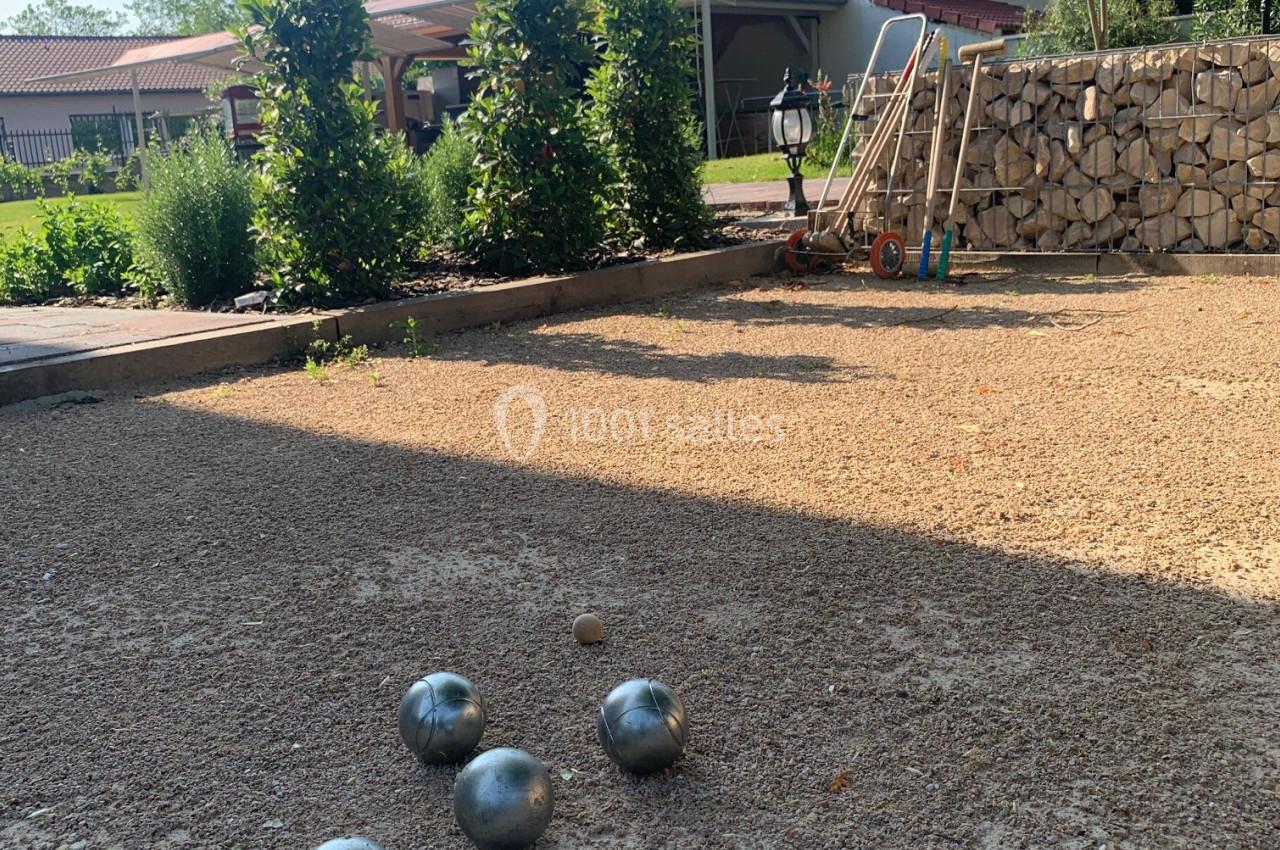 Terrain de pétanque avec des boules en métal et un cochonnet, entouré de végétation et d'une maison en arrière-plan.