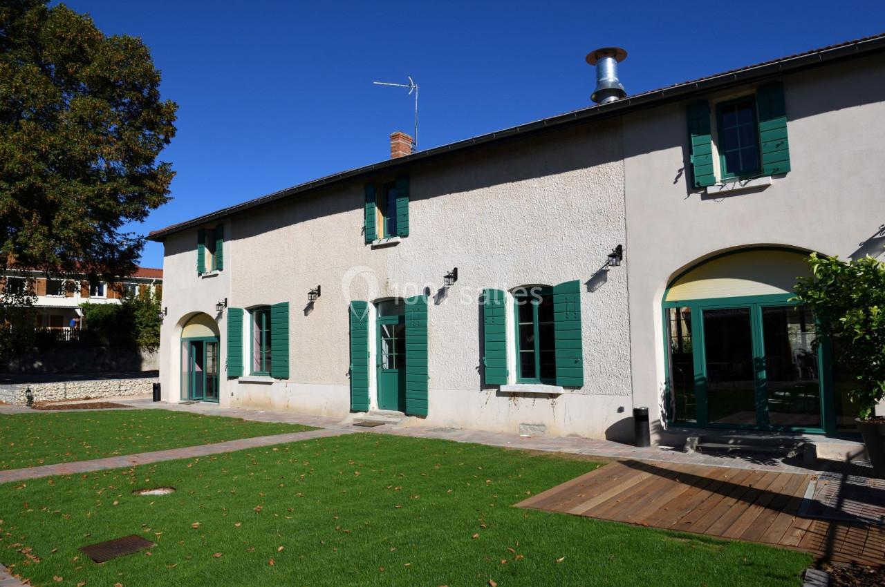 Façade d'une maison avec volets verts, pelouse et terrasse en bois sous un ciel bleu clair.