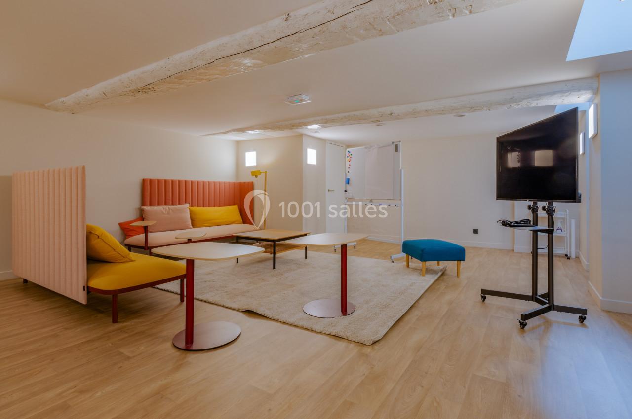 Salle lumineuse avec parquet, mobilier coloré, tables basses, canapé, écran sur pied et poutres apparentes au plafond.