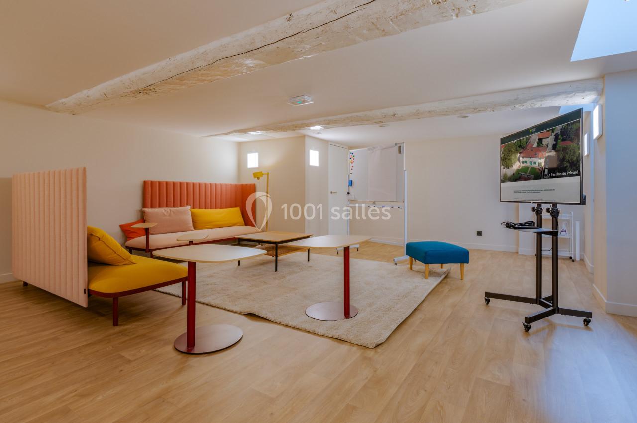 Salle lumineuse avec parquet, mobilier coloré, poutres apparentes et écran sur pied dans un espace aménagé.