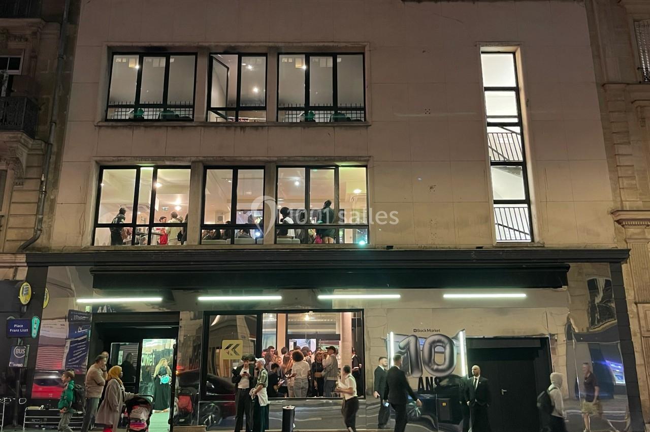 Façade d'un bâtiment moderne éclairé, avec des passants et des invités visibles à l'intérieur et à l'extérieur.