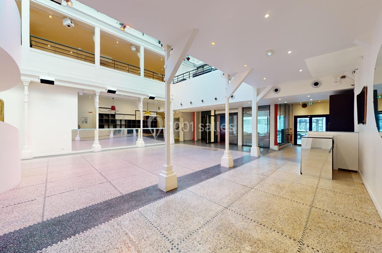 Intérieur d'un espace lumineux avec sol en terrazzo, colonnes blanches, mezzanine et grandes baies vitrées.