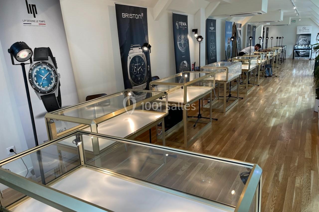 Salle d'exposition lumineuse avec vitrines en verre vides alignées sur un sol en bois, affiches de montres au mur.