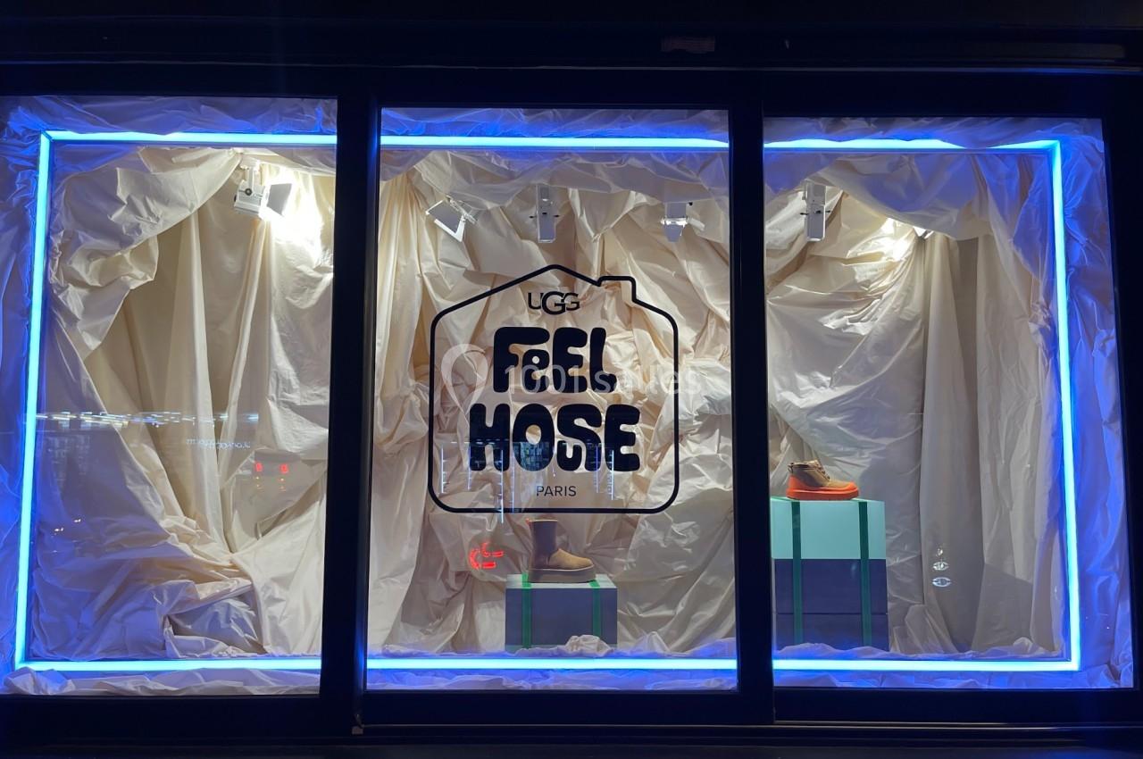 Vitrine éclairée avec des chaussures exposées sur des socles, entourées de rideaux beige et encadrée de néons bleus.