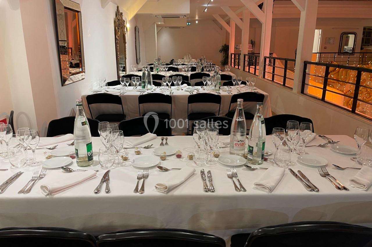 Salle de réception avec tables dressées, nappes blanches, couverts, verres et bouteilles d'eau, éclairage chaleureux.