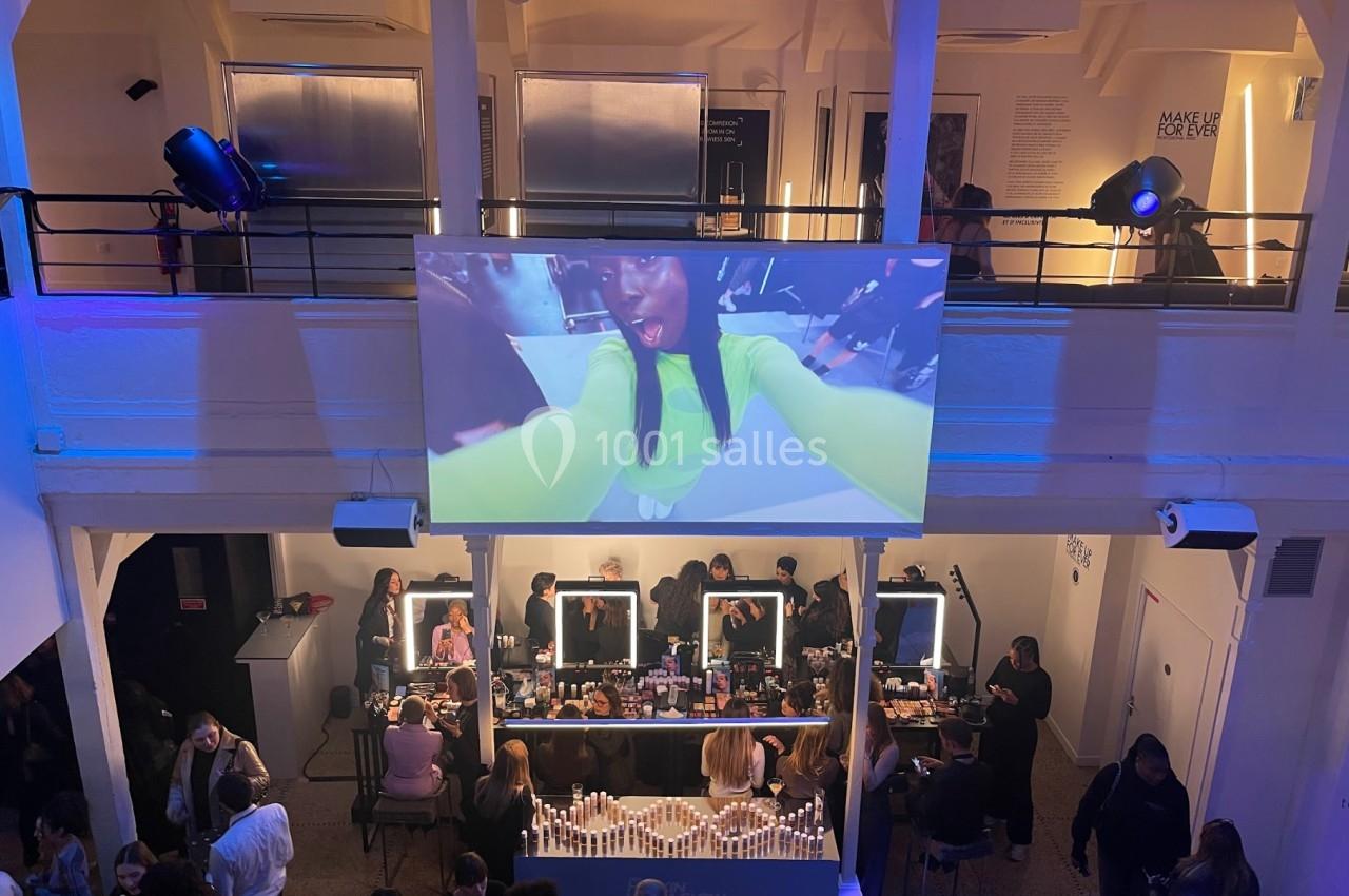 Vue d'un événement avec un écran géant affichant une femme en gros plan et des participants autour de tables de maquillage.