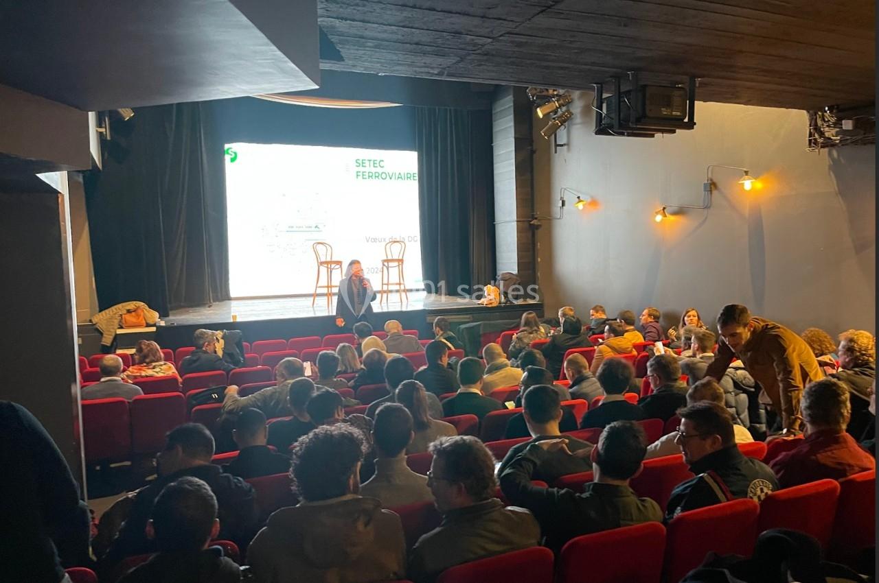 Salle de spectacle avec des spectateurs assis, une scène éclairée et une présentation en cours.